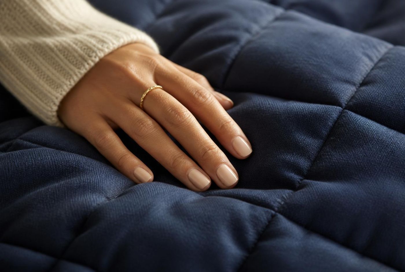 Nærbillede af en hånd der hviler på en mørkeblå quiltet tyngdedyne i blødt vinduslys — fokus på quilting og fint vævet stof